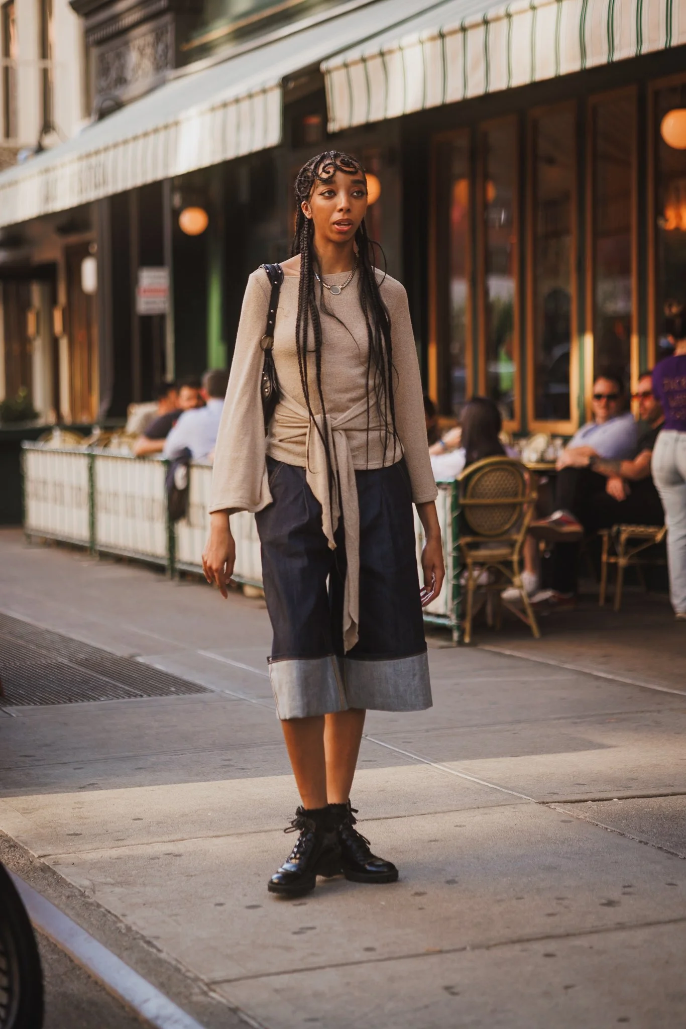 A young woman walking on a city sidewalk in front of a restaurant or cafe with outdoor seating. She has long braided hair, is wearing a beige sweater tied at the waist, a long mixed fabric skirt, and black combat boots. She has a black shoulder bag a