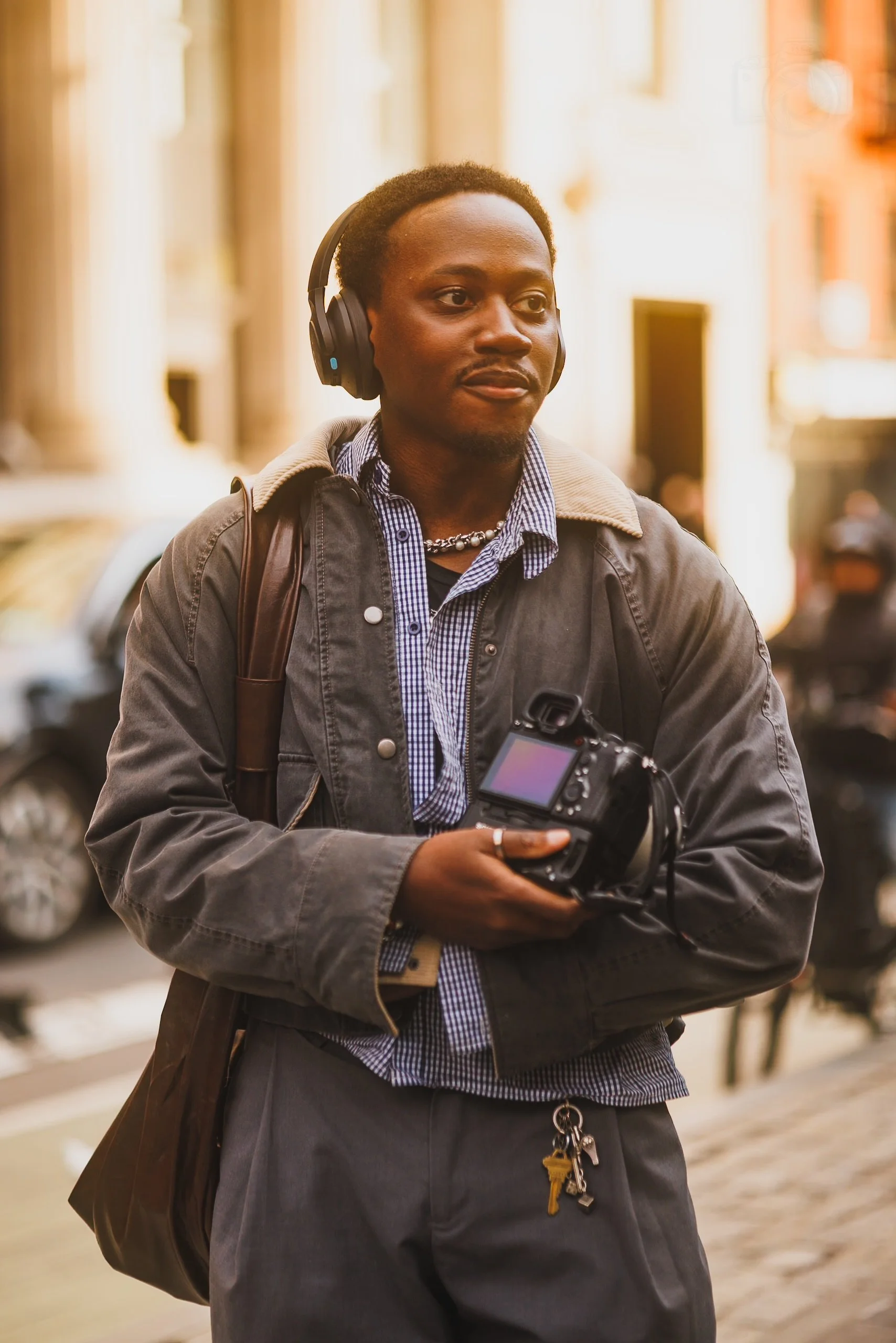 A man walking outdoors in a city, wearing headphones, a gray jacket, a plaid shirt, and carrying a camera and a bag.