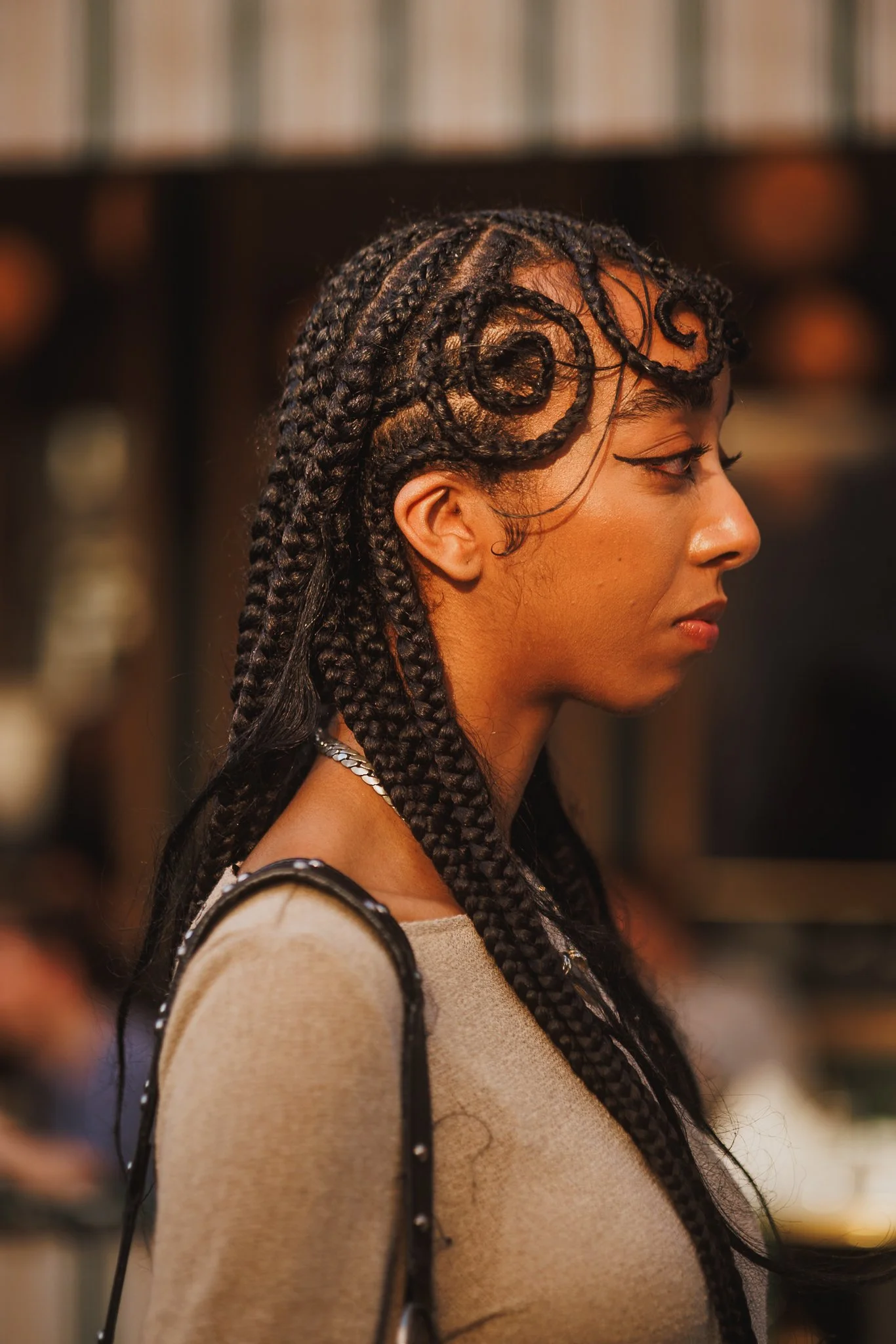 Close-up of a woman's profile with braided hair styled in intricate swirls, wearing makeup with winged eyeliner, and dressed in a beige top with thin straps, against a blurred background.