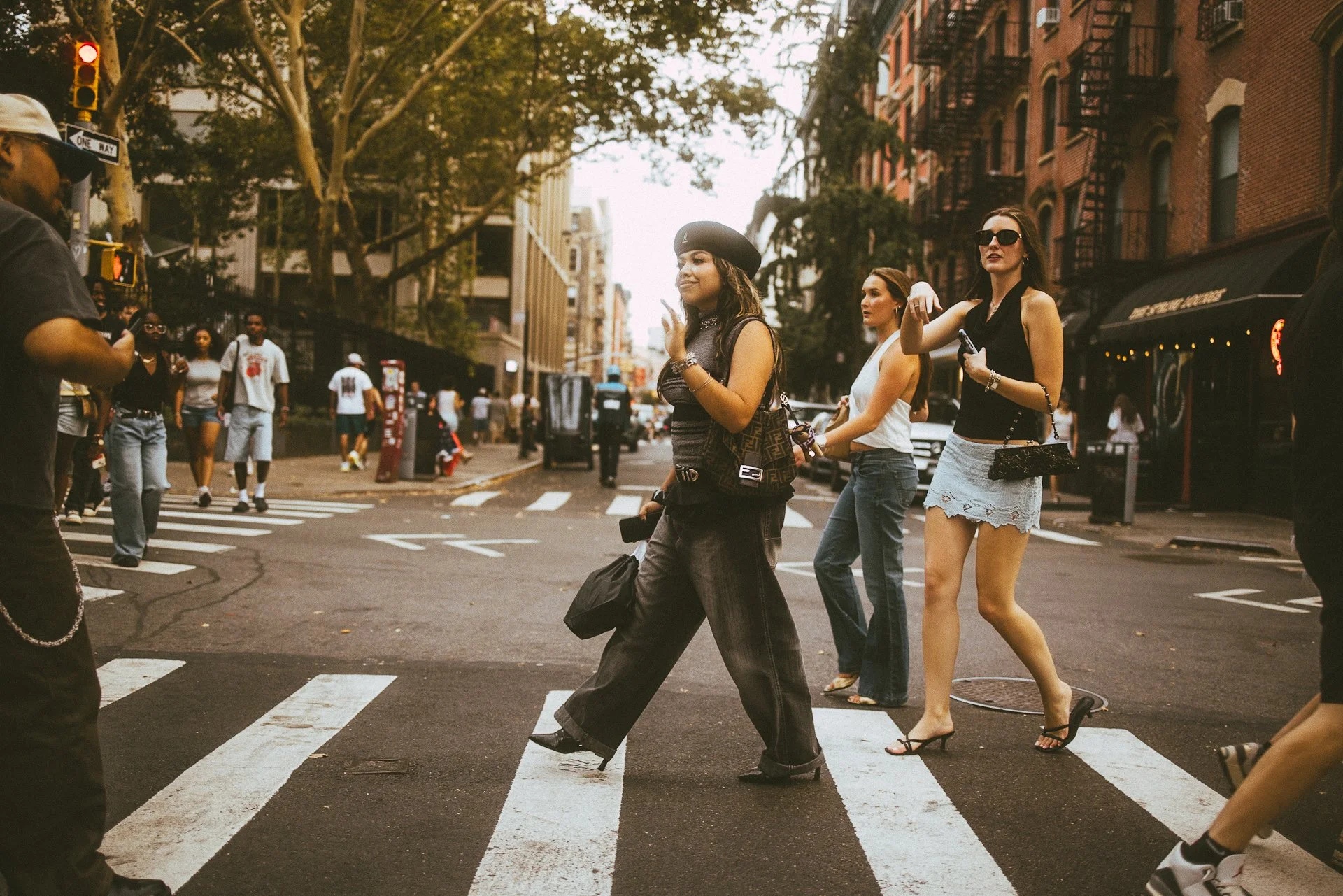 People crossing a busy city street at a crosswalk during daytime, with buildings, trees, and storefronts in the background.