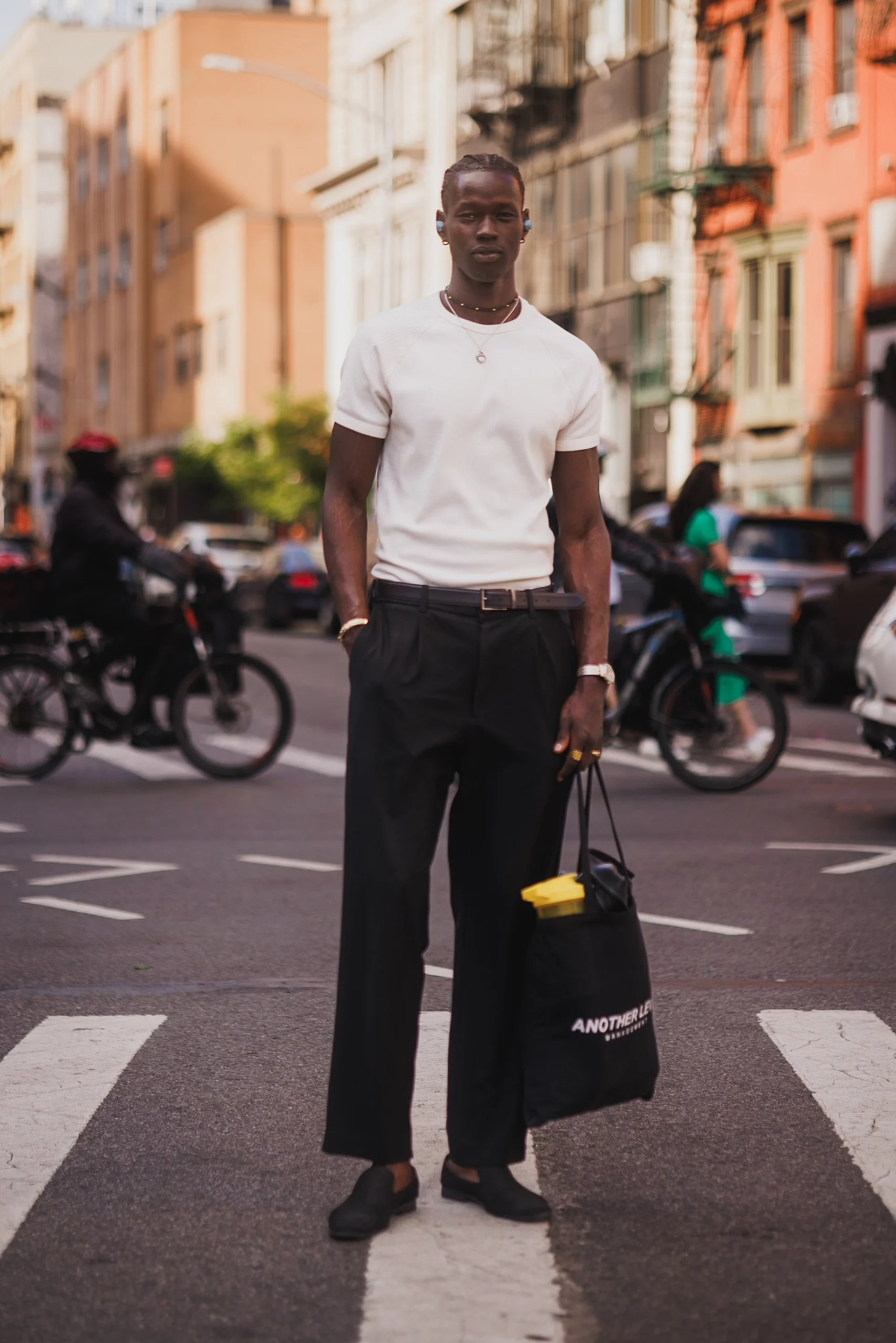 A young man with dark skin and short hair stands on a crosswalk in an urban area during daytime. He is wearing a white t-shirt, black pants, and black shoes, and has a necklace and earrings. He is holding a black bag with yellow and white text that s