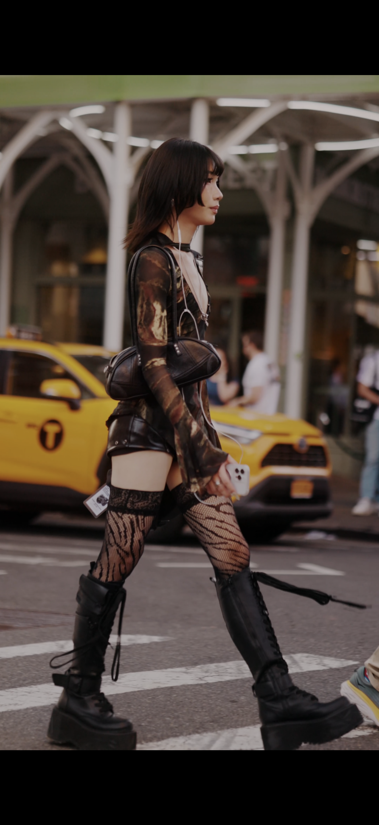 A young woman walking across a city street during daytime, holding a smartphone, dressed in a sheer patterned outfit, and wearing high platform black boots, with taxis and pedestrians in the background.