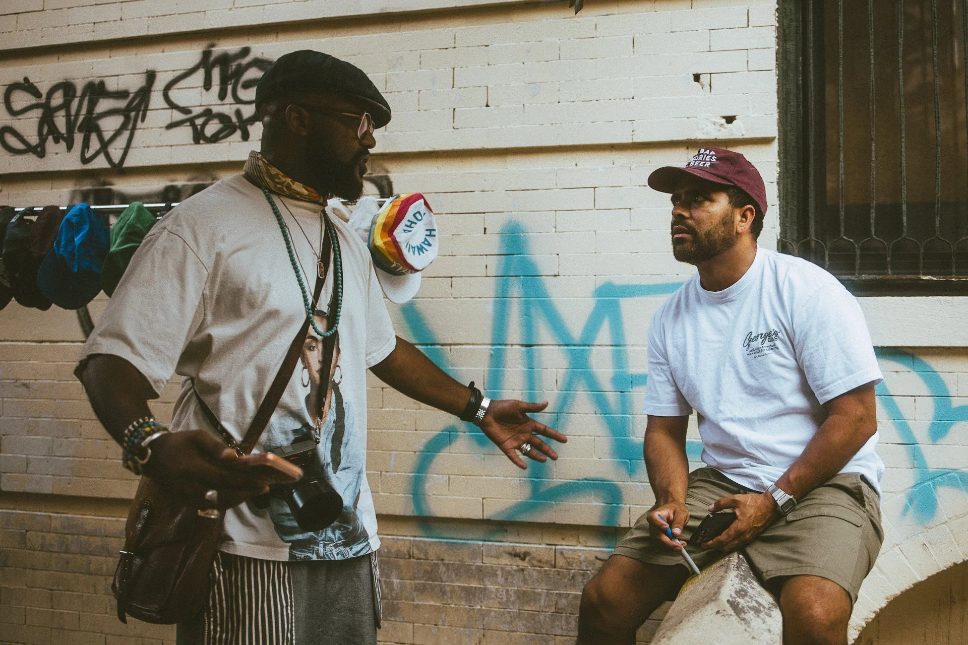 Two men are having a conversation against a white brick wall with graffiti. One man is standing, wearing a flat cap, glasses, a graphic t-shirt, and striped pants, holding a camera. The other man is sitting on a concrete ledge, wearing a maroon cap, 