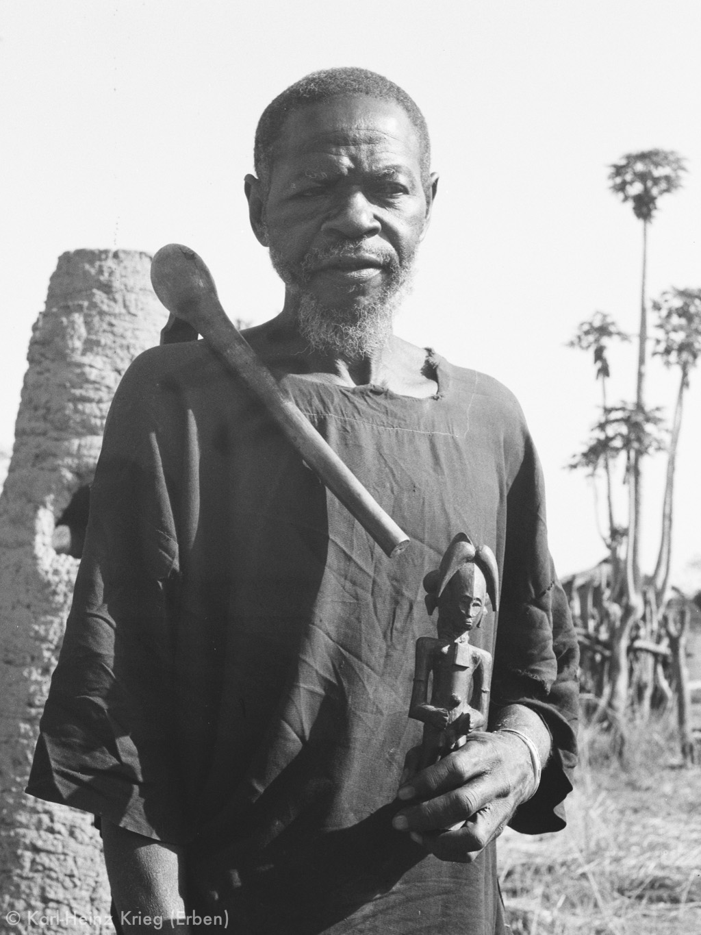 Kolotielema Dagnogo with a figure that he carved. On his right shoulder he carries an adze , the symbol of a wood carver. Behind him are old furnaces used to smelt iron in the past. Photo: Karl-Heinz Krieg, Poundiou (Region of Boundiali), 1974