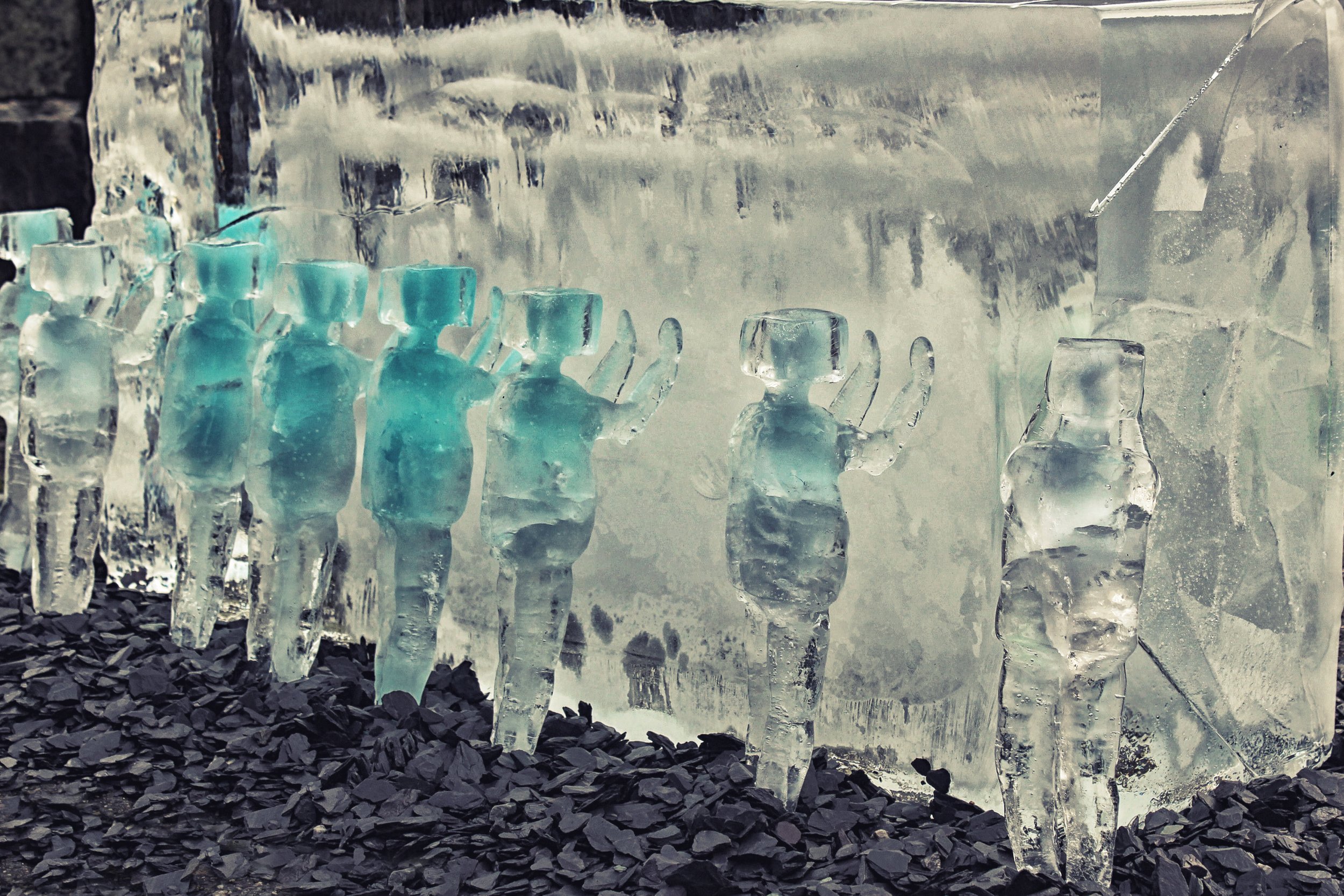 Small ice sculptures of human figures with turquoise hues in a line against a large ice block backdrop, set on a rocky surface.