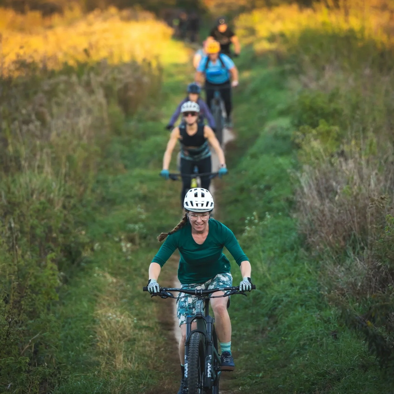 Group of people riding mountain bikes on a forest trail surrounded by tall trees and bushes.