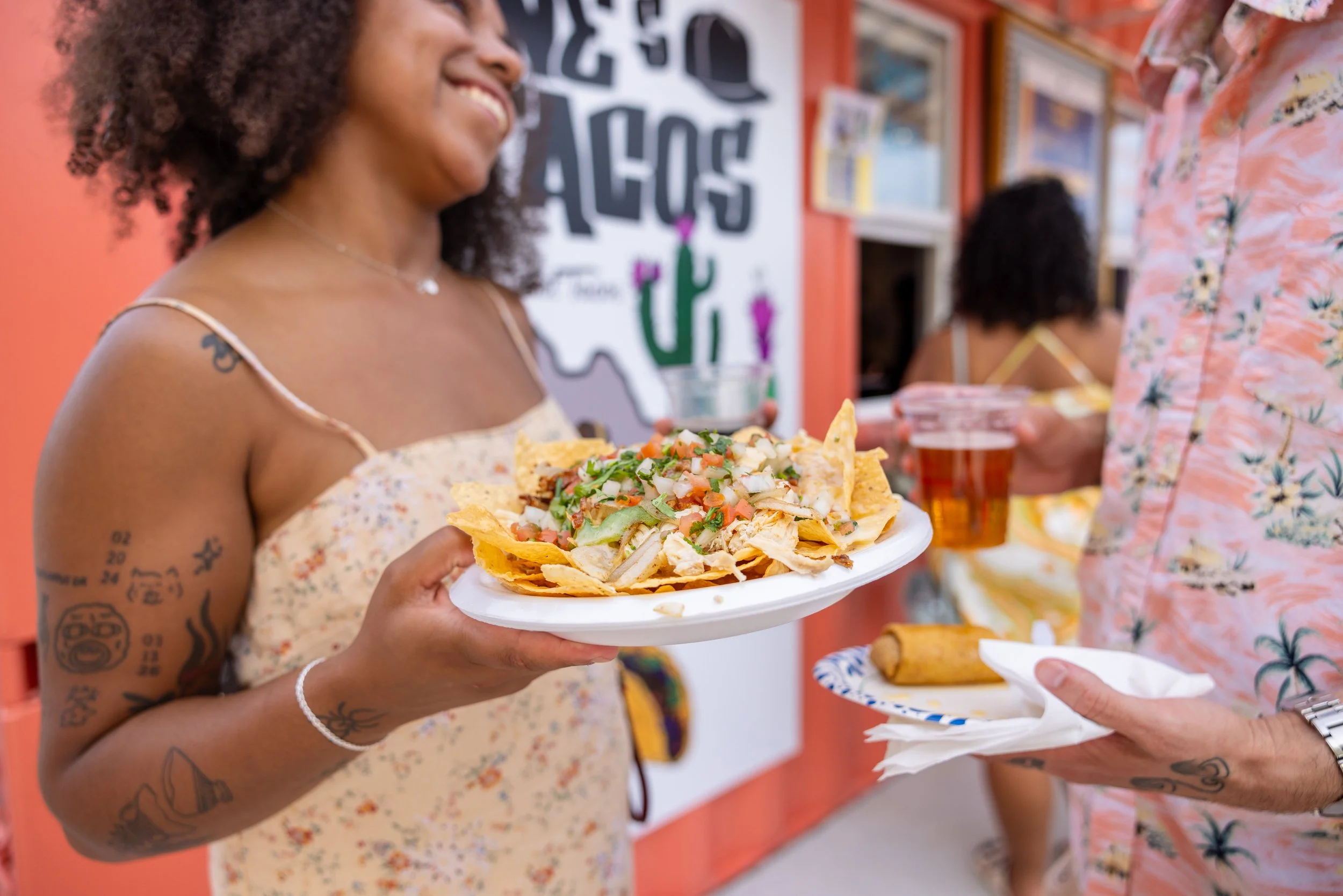 A woman holding a plate of nachos with cheese, tomatoes, and green herbs, smiling at a food stand. Other people holding drinks are visible in the background.