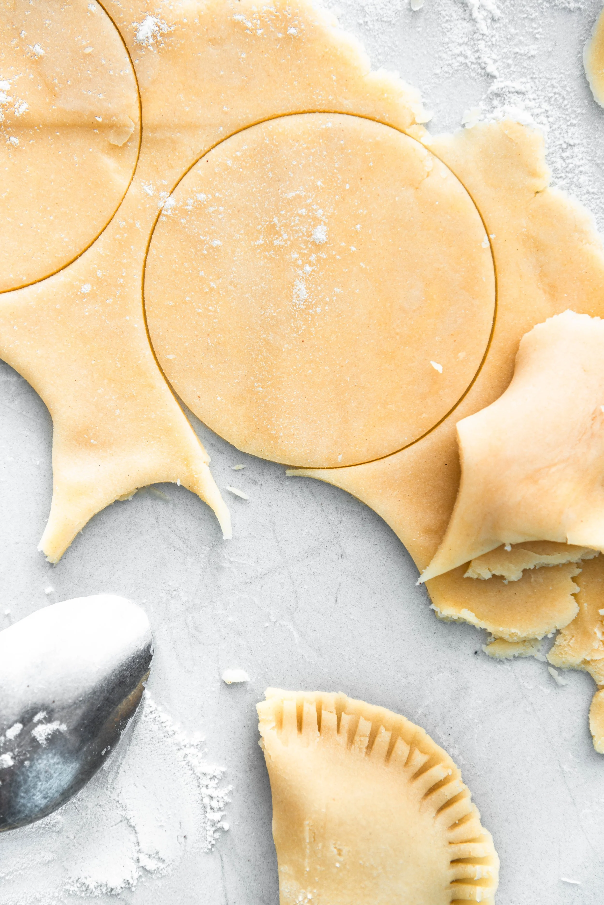 Unrolled pie crust dough with round cookie cutter, cut circles and trimmed edges on a floured surface, with a pastry cutter in the bottom left.