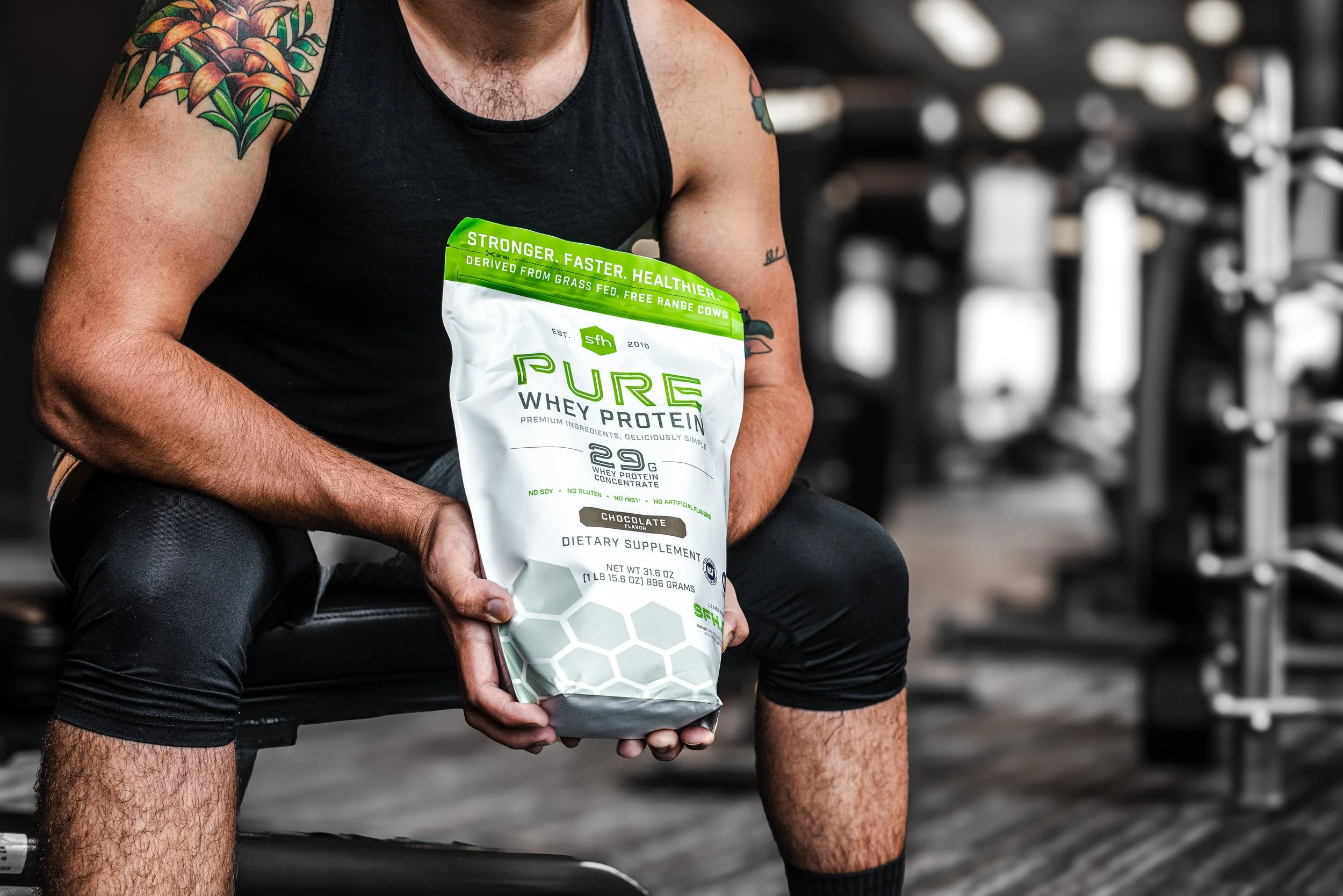 A man sitting on a gym bench, holding a large bag of 'Pure Whey Protein' in front of him. He is wearing a black tank top with tattoos on his arms, black athletic leggings, and black socks, with gym equipment visible in the background.