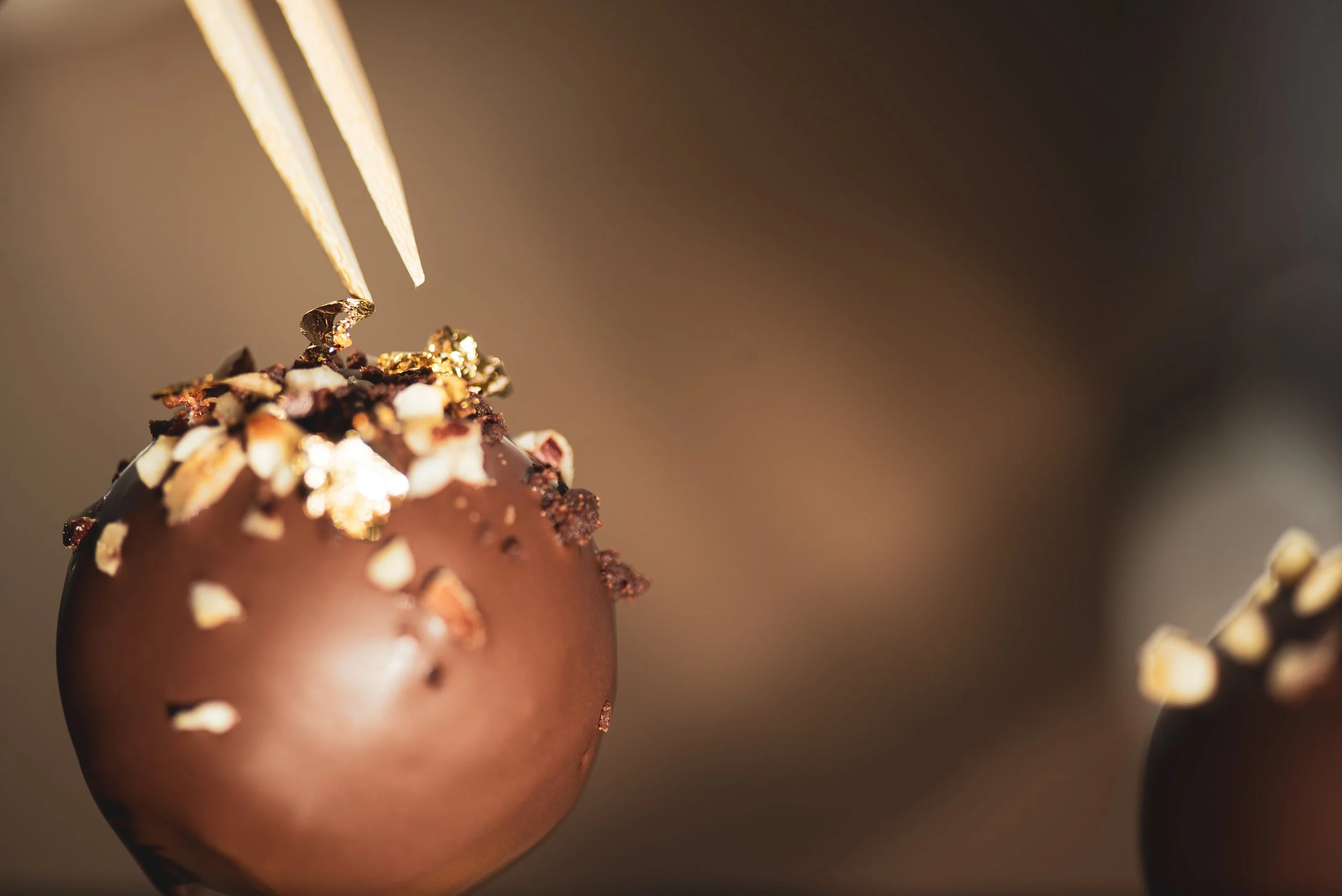 Close-up of a chocolate truffle topped with chopped nuts and edible gold leaf, with a gold fork inserting into it.
