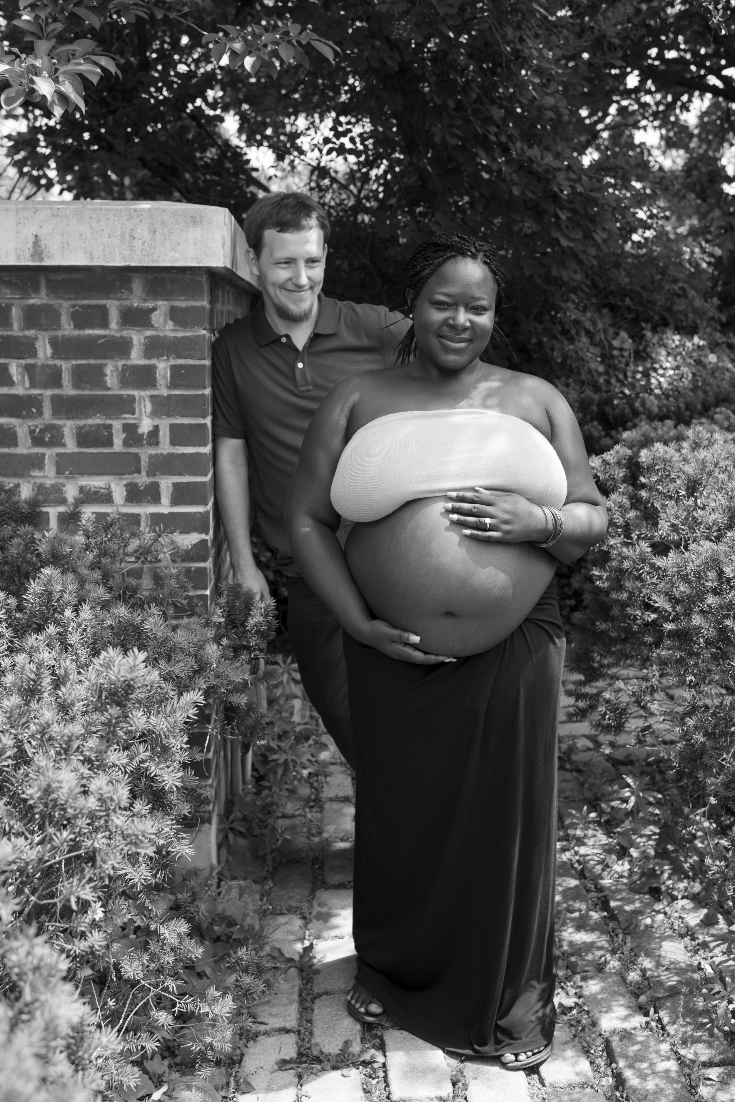 Pregnant woman standing outdoors, holding her belly, with a man standing behind her leaning on a brick wall. Black and white photo.