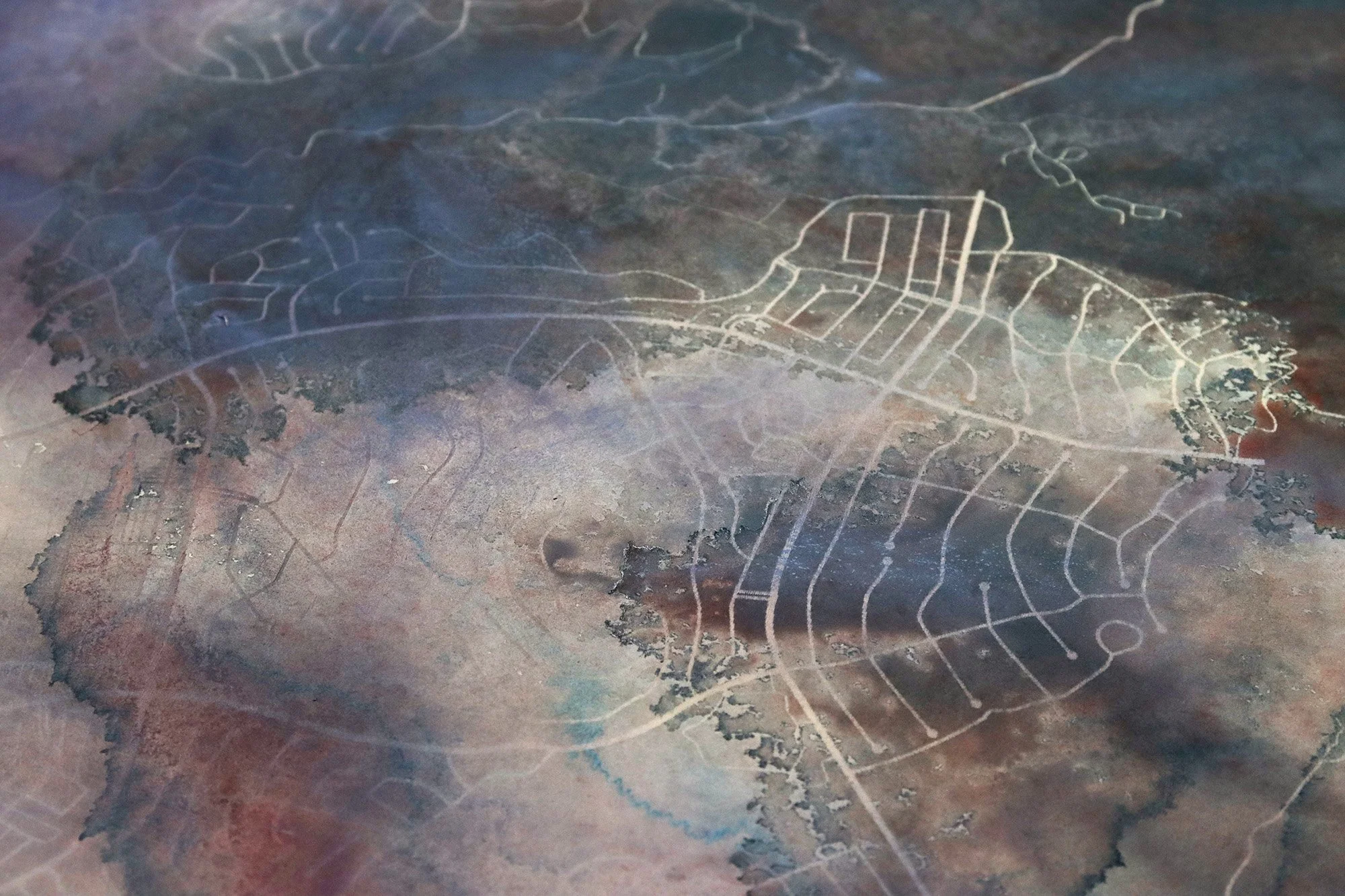 Pale laser-engraved street and plot geometries emerging through layered blue-grey, mauve and ochre acrylic washes on paper.