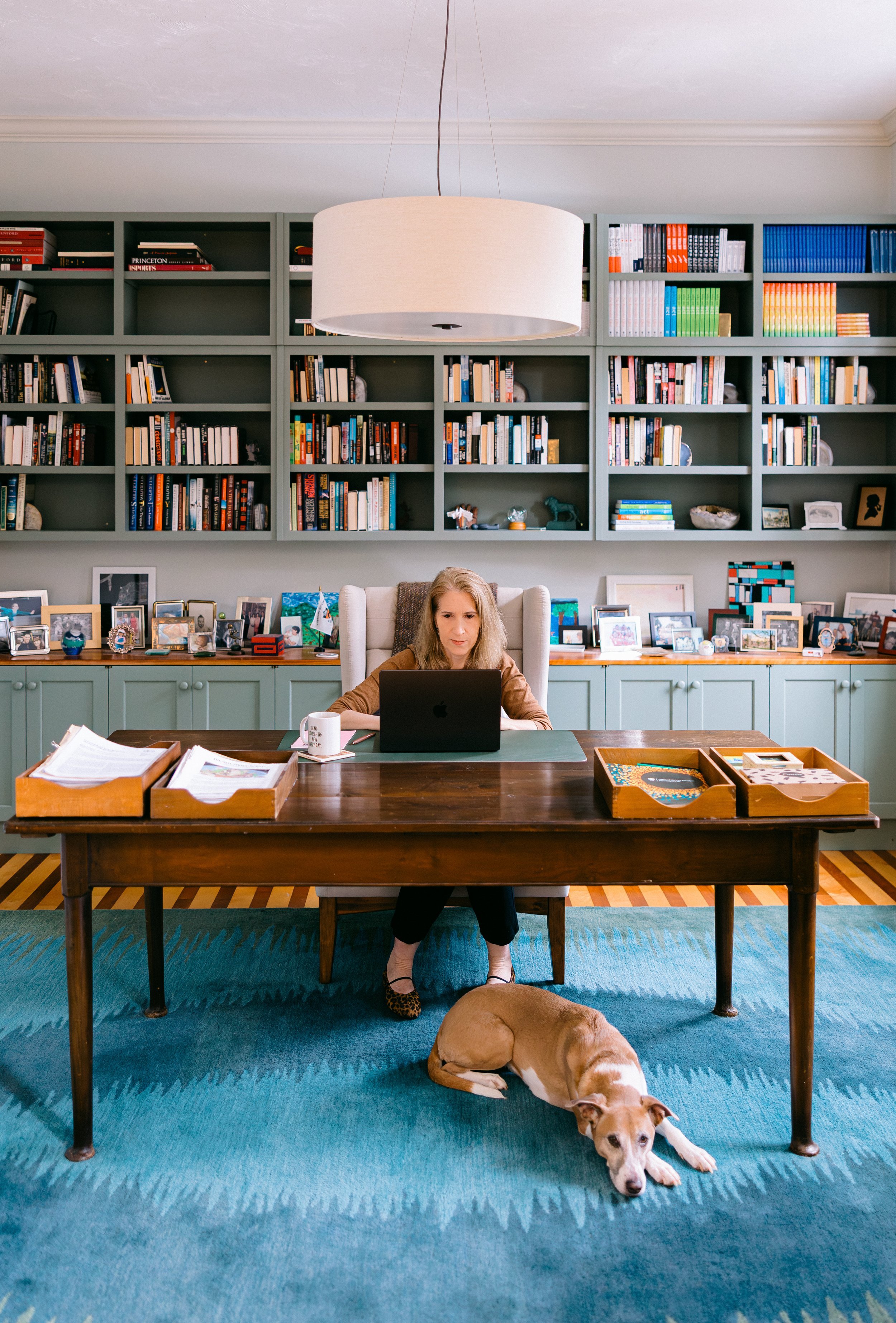 Catherine sits at a desk with her laptop, a dog laying at her feet