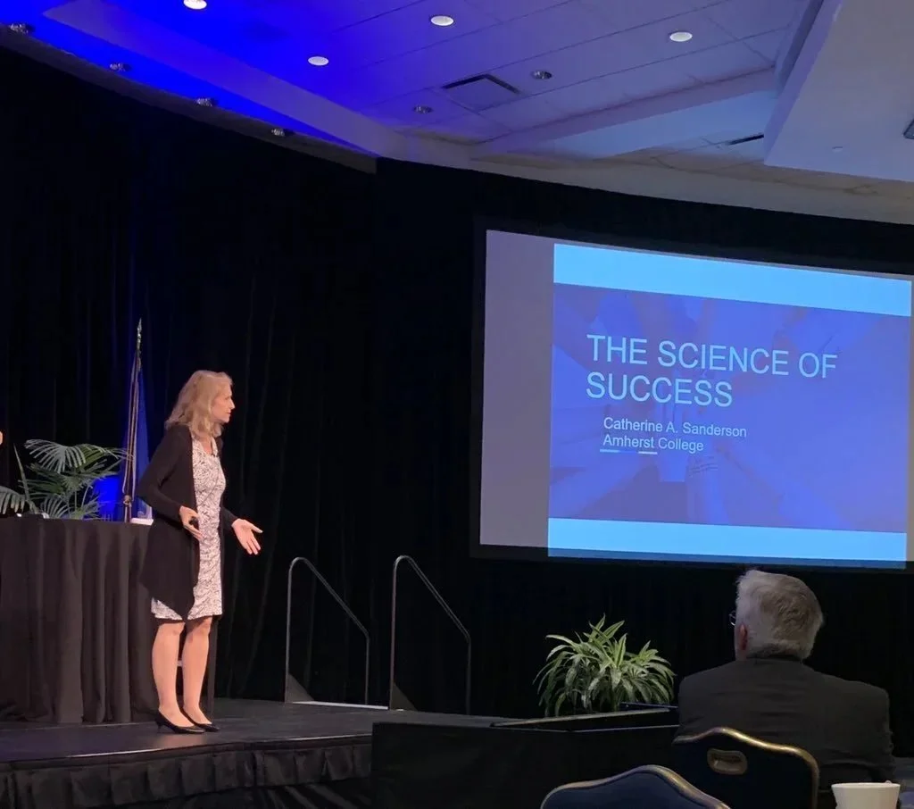 Catherine stands onstage with a presentation titled, "The Science of Success"