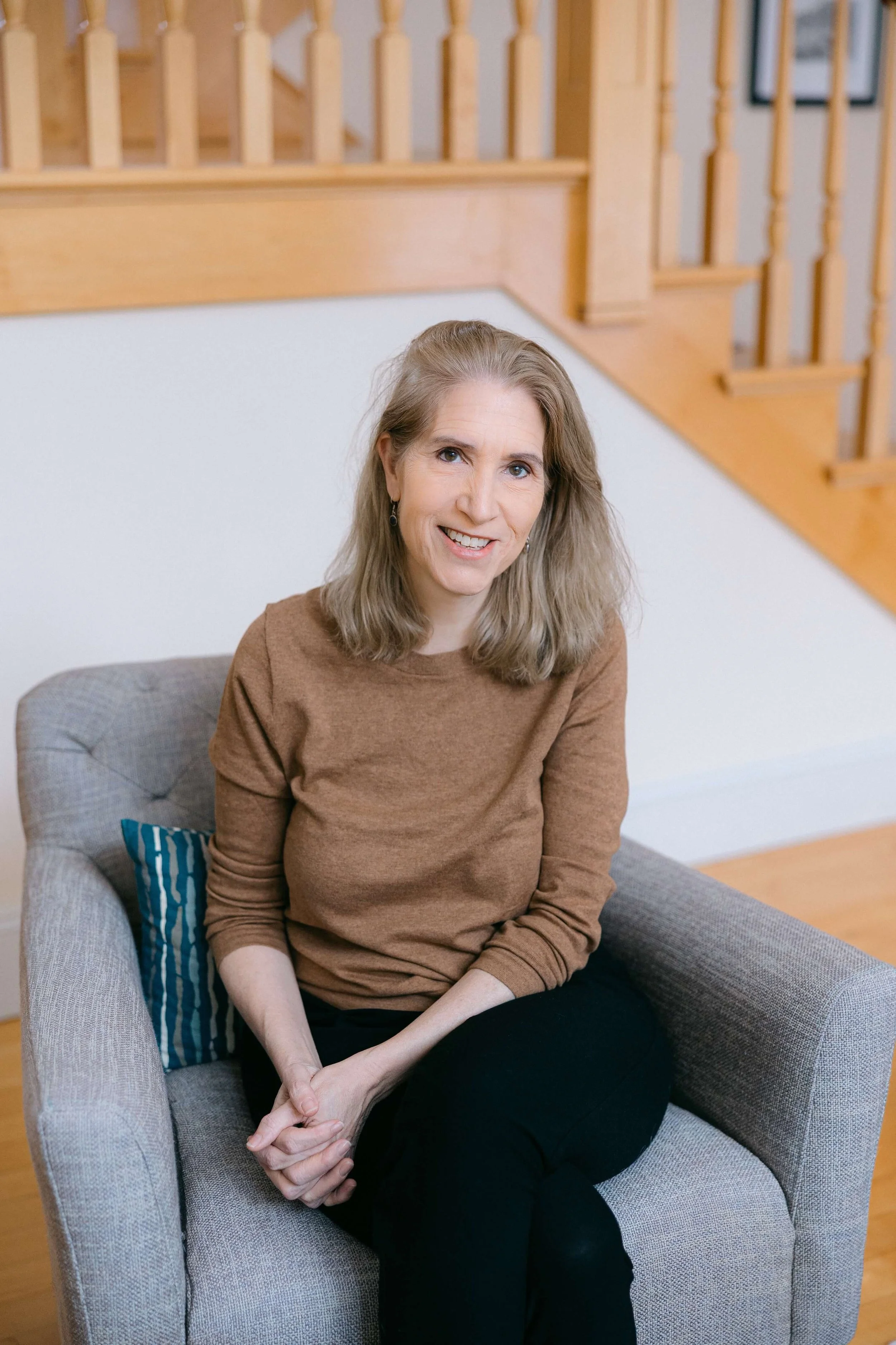 Catherine smiles in a brown sweater, sitting on a gray armchair
