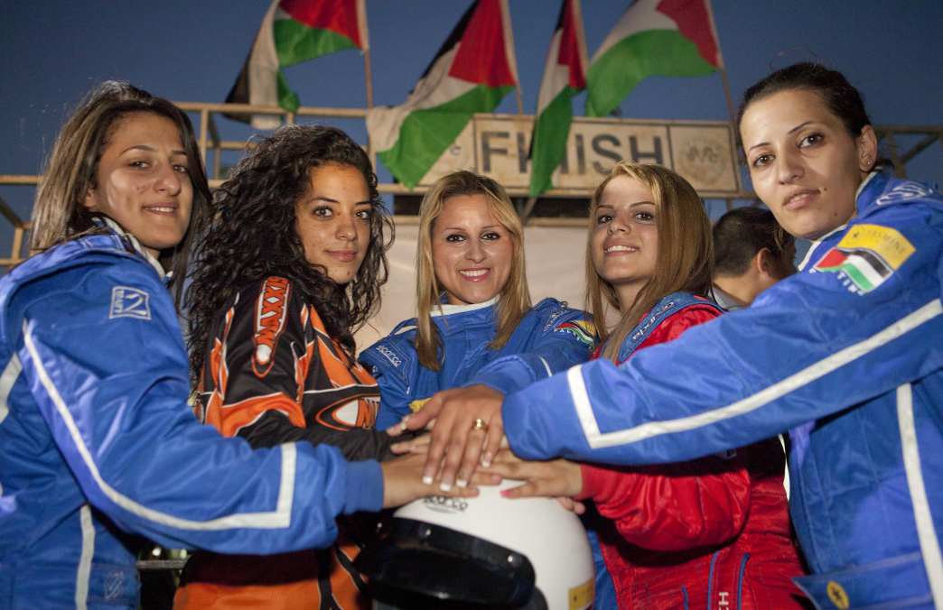 five-women-in-race-car-jackets-smiling-to-the-cmarea-with-their-hands-on-one-helmet-in-the-middle-of-them.PNG
