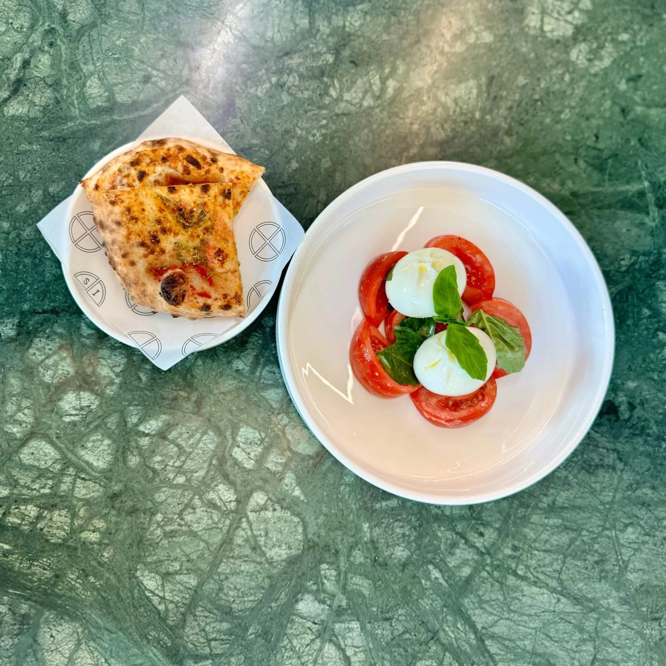Insalata Caprese: A plate with tomato and mozzarella salad garnished with basil leaves and a bowl with two slices of pizza on a green marble table. Two sliced of housemade foccacia sit on the side.
