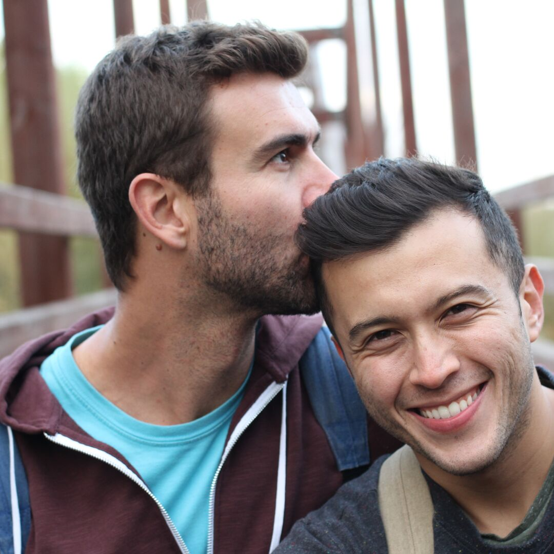 Gay male couple kissing head on bridge.png