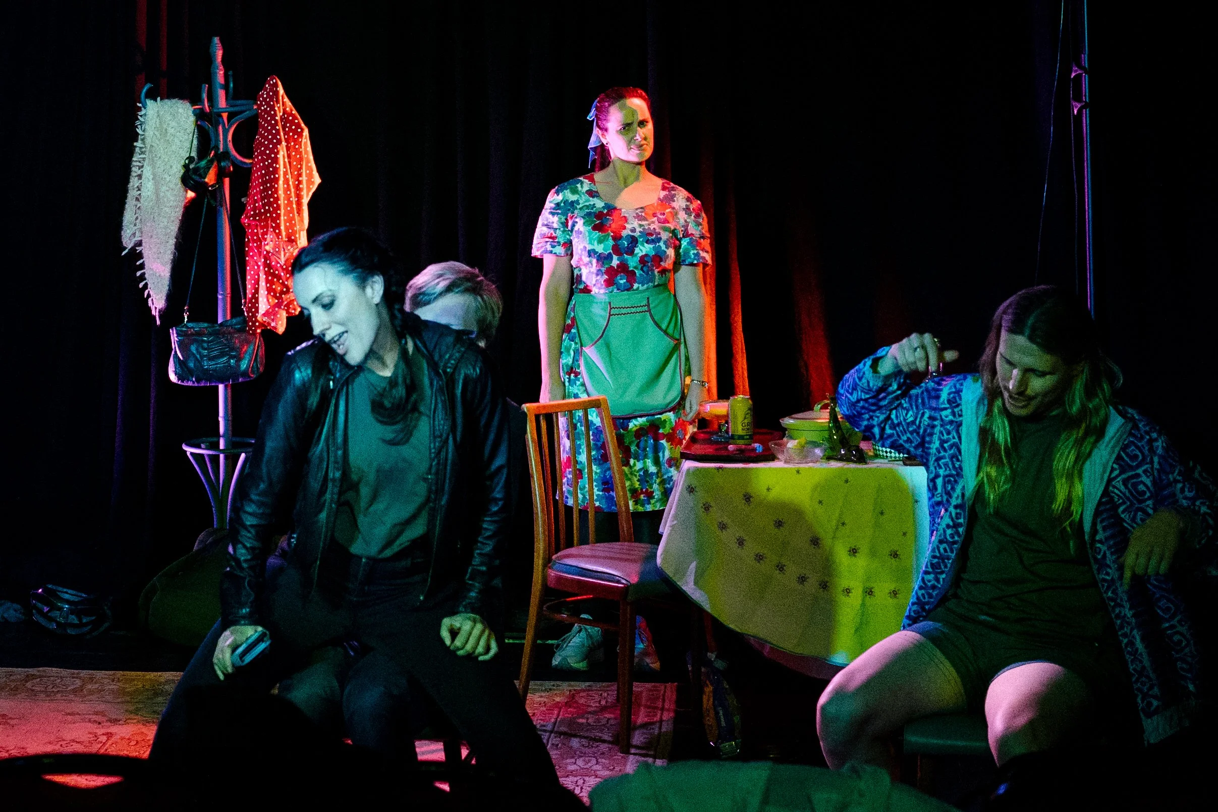 A group of people dancing with colorful lighting, a woman standing in the background near a clothing rack with various garments, and a girl with dark hair dancing in front