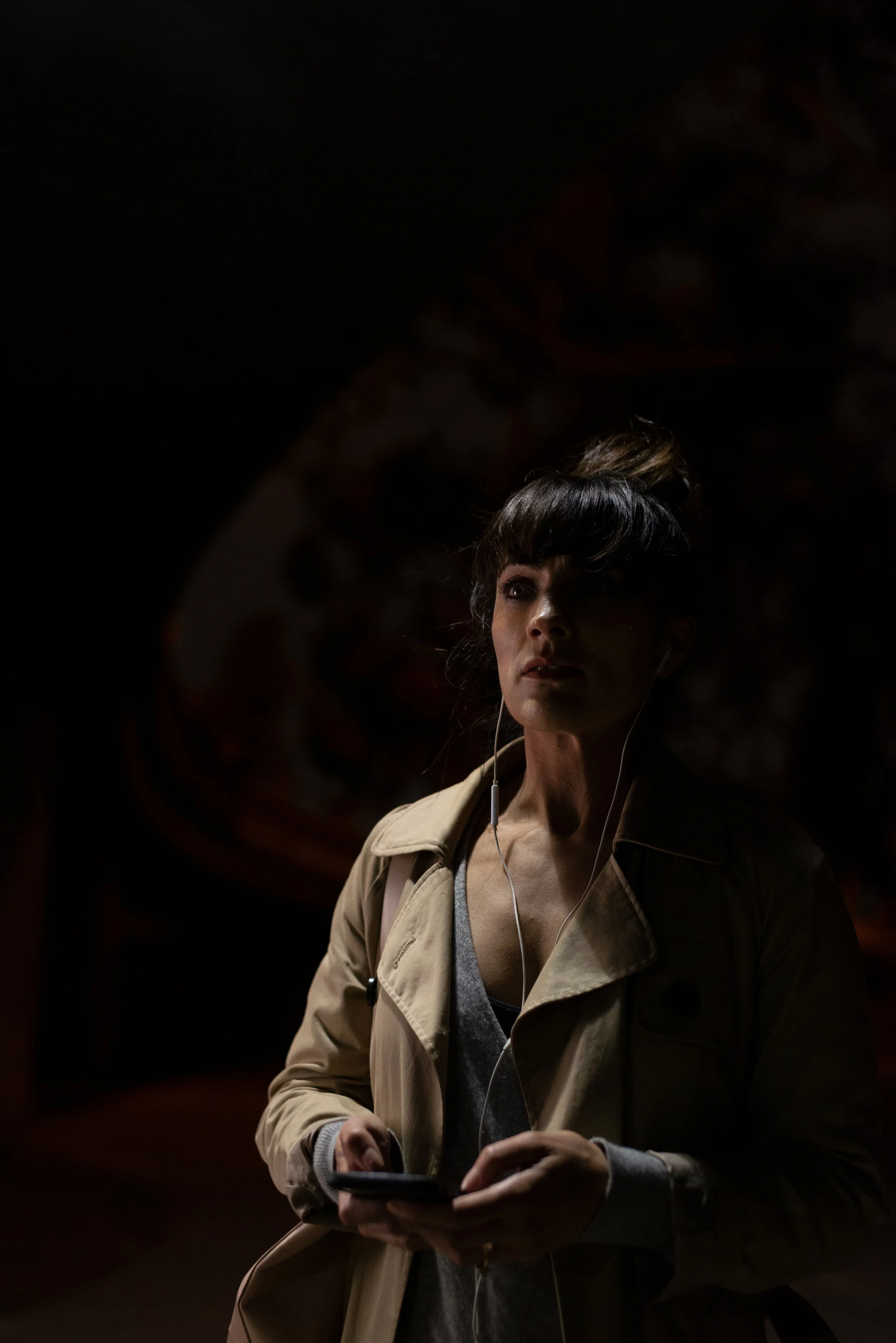 A woman with dark hair styled in a bun, wearing a beige jacket and gray sweater, holding a smartphone, with earphones in, walking in a dimly lit environment.