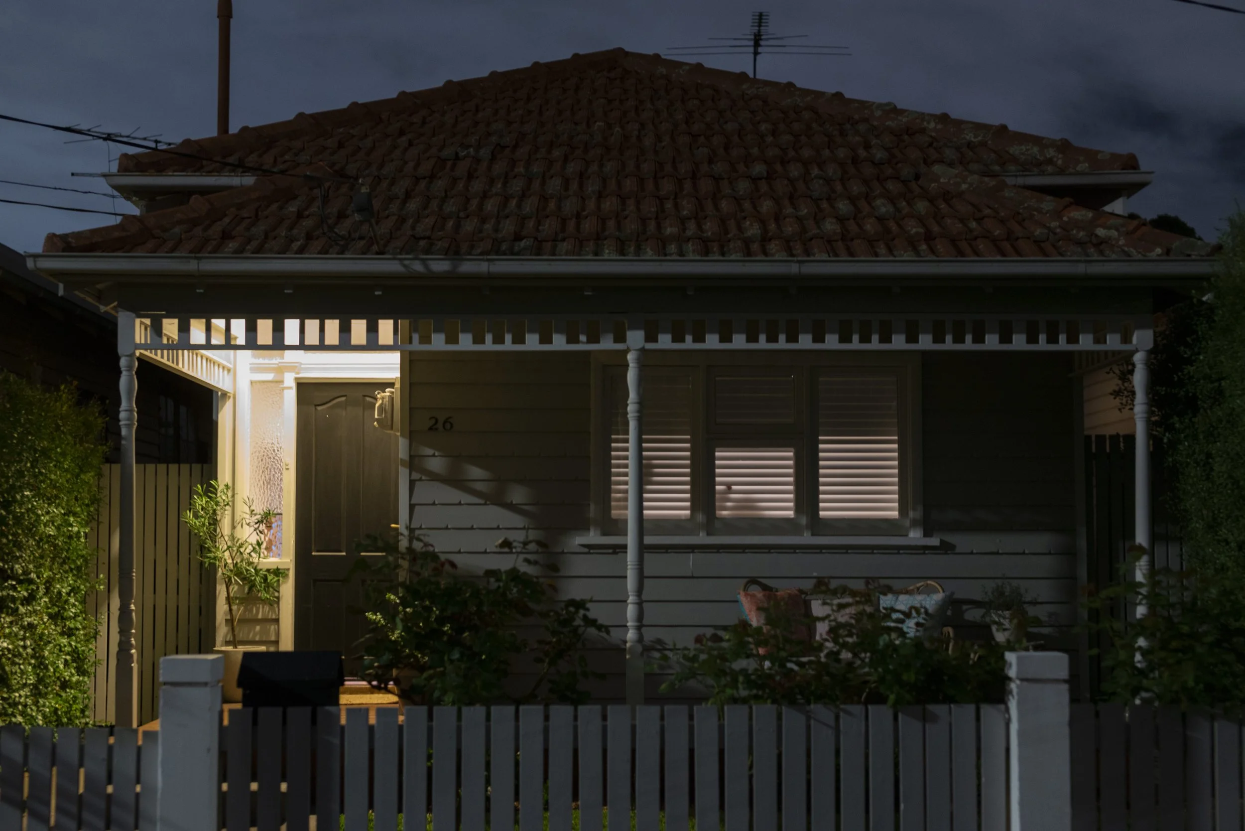 A house with a tiled roof, lit from inside, taken at dusk or night. The house has a porch with white columns, a front door, a window, and a white picket fence surrounding it. There are trees and bushes outside.