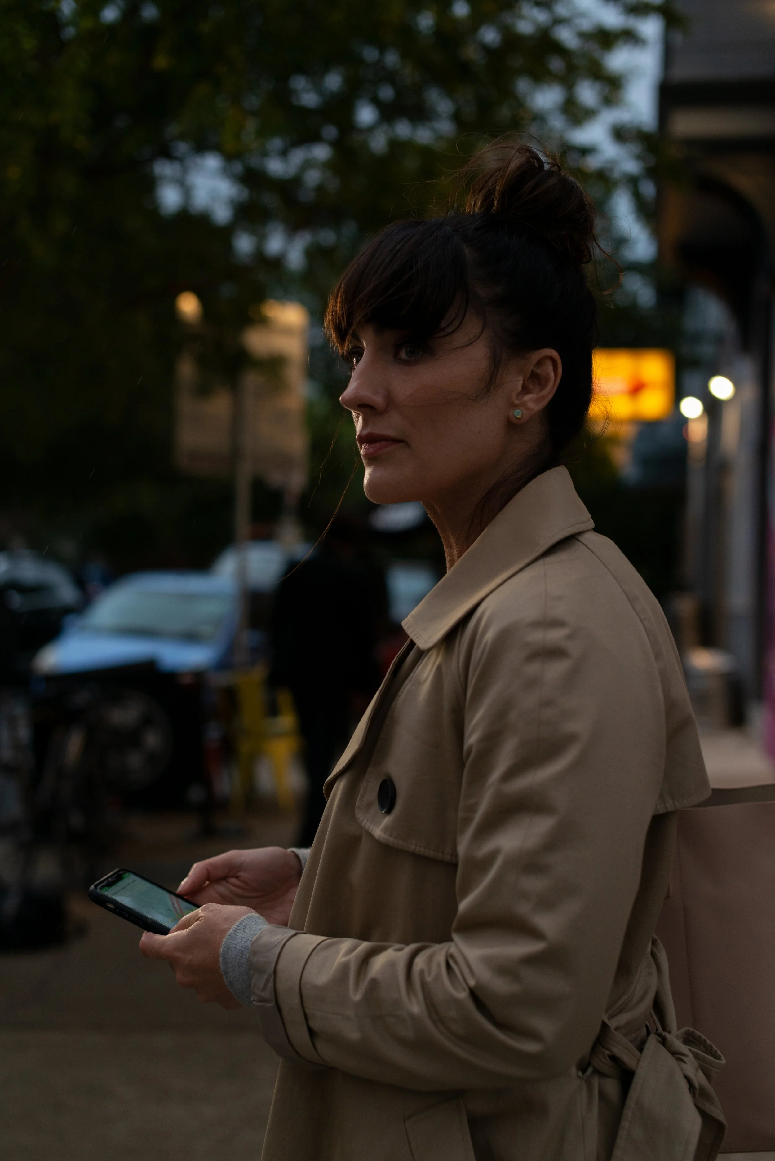 A woman with dark hair tied up in a bun, wearing a tan trench coat, standing outdoors at dusk and looking at her phone.