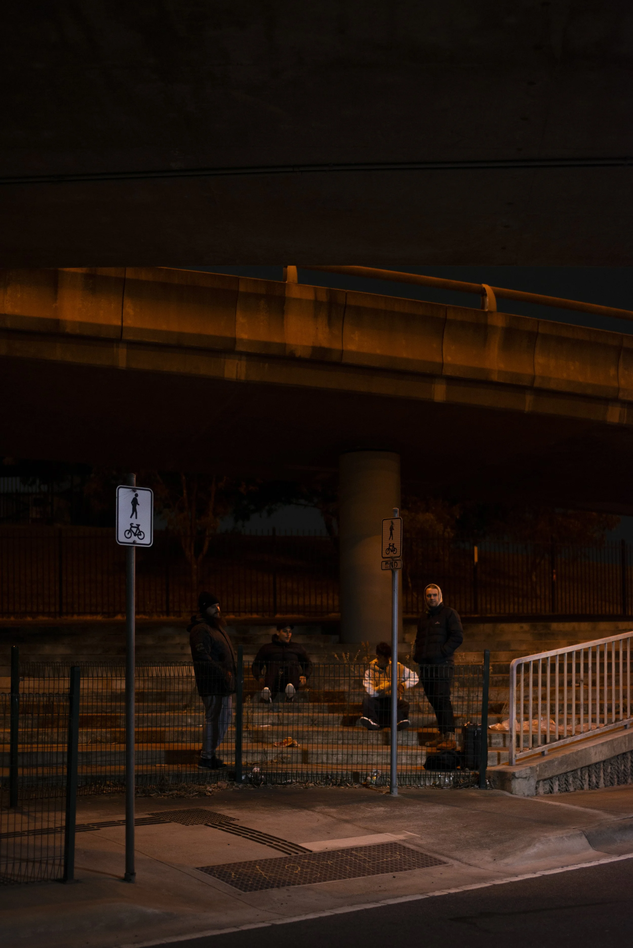Nighttime scene under an overpass with a group of people standing and sitting. There are signs indicating a pedestrian and bicycle pathway, and a small fenced area in the foreground.