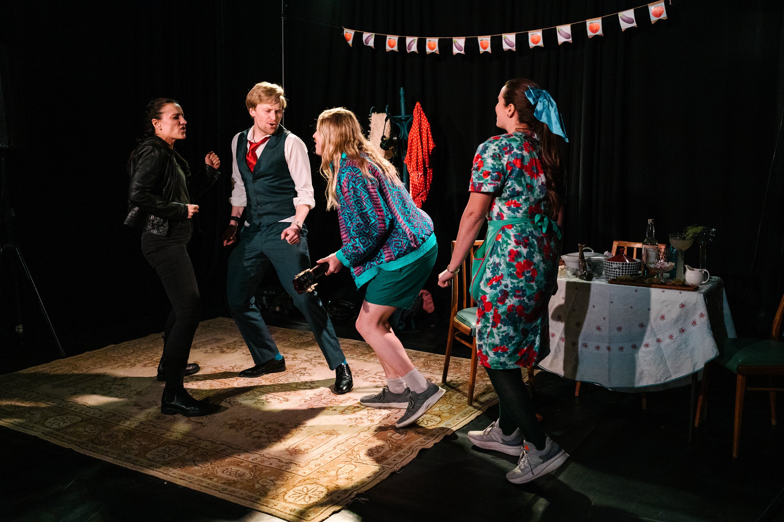 Four actors are performing on a stage with a black background and a banner of fruit and vegetable flags overhead. They appear to be in a lively scene Two women, one in a leather jacket and the other in a floral dress with a blue headscarf, and two men, one in a vest with a red necktie and the other holding a camera, are engaged in the scene. There is a table with various dishes and bottles on the right side.