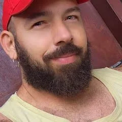 Close-up of a man with a beard and mustache wearing a red cap and yellow shirt, smiling at the camera.