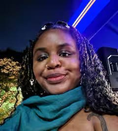 A young woman with braided hair, earrings, and a nose ring, smiling outdoors at night, with blue lights in the background.