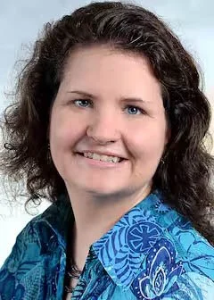 Portrait of a woman with curly brown hair, wearing a blue patterned shirt, smiling at the camera.