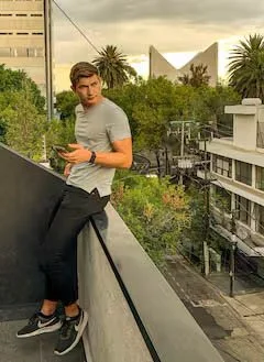 Young man sitting on a balcony railing, holding a phone, with palm trees and modern buildings in the background.