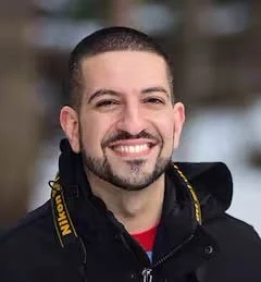 A man with short dark hair and a beard smiling at the camera, wearing a black jacket with yellow accents.