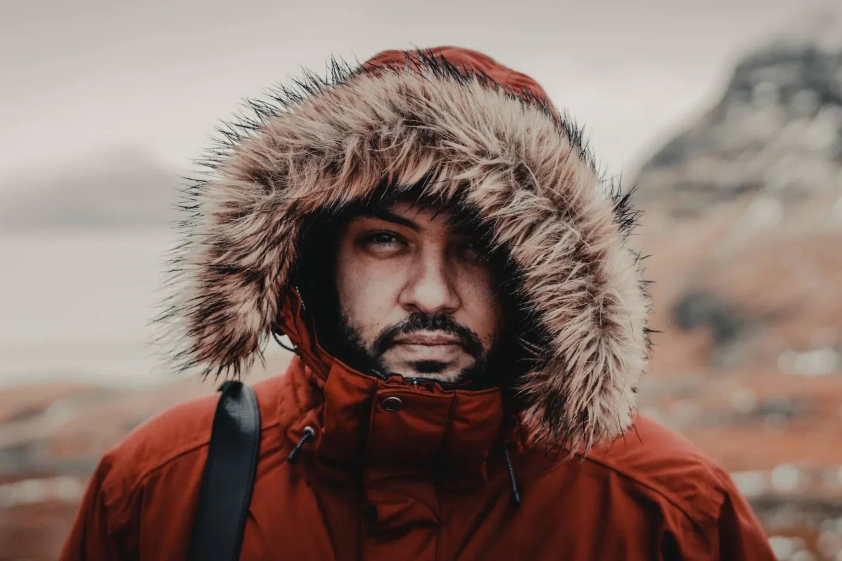 A man with a beard wearing a red winter coat with a fur-lined hood outdoors in a cold environment.