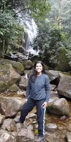 A woman standing on rocks in a forested area with a waterfall in the background.