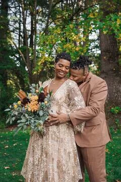 A couple dressed in formal attire standing outdoors among trees, smiling and embracing each other, with the woman holding a bouquet of flowers.