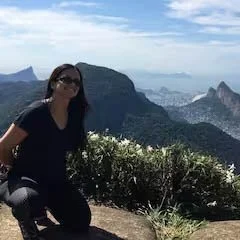 A woman wearing sunglasses and a dark shirt, sitting on a rock with a scenic mountain landscape and cloudy sky in the background.