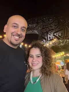 A man and a woman smiling outdoors at night, with decorative lights and structures in the background.
