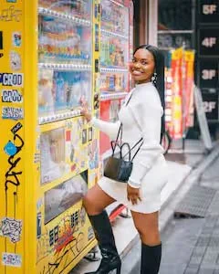 A woman in a white outfit and black boots smiling while buying snacks from a vending machine on a city street.