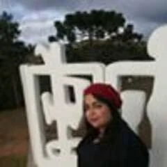 A woman wearing a red headscarf standing in front of a white decorative fence outdoors with trees and dark clouds in the background.