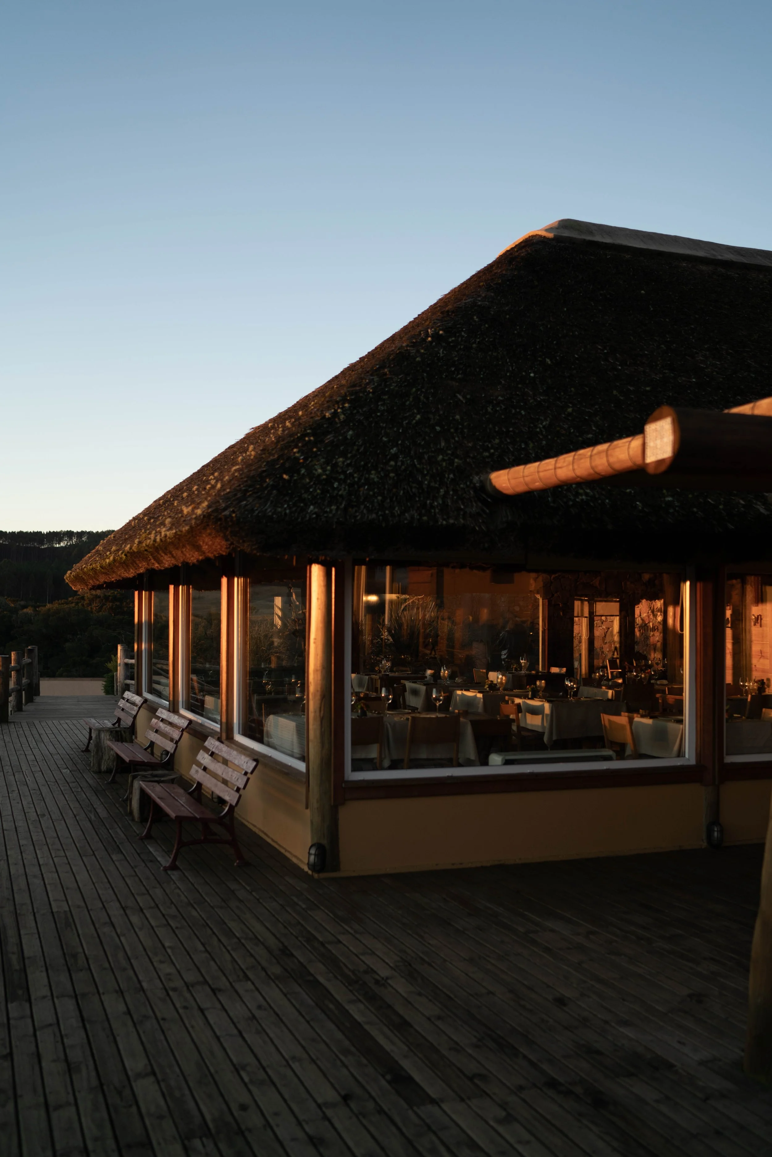 An outdoor restaurant with a thatched roof, large windows, and wooden benches on a deck, with sunset lighting and a clear sky.