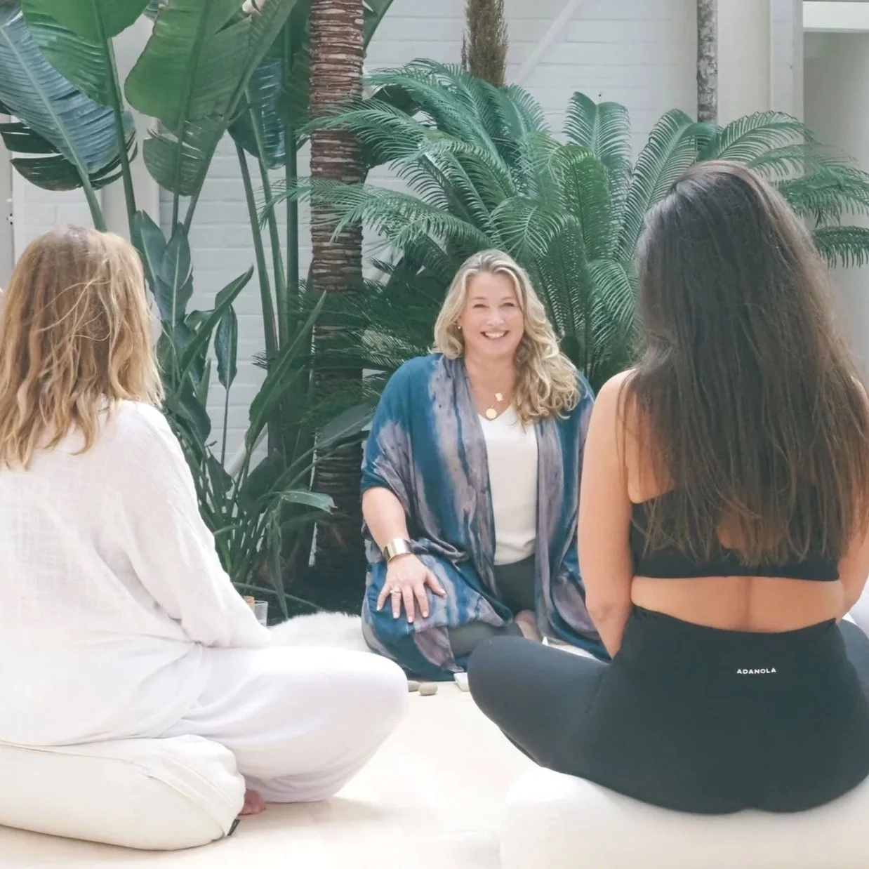 Three women sitting on a white floor surrounded by large green tropical plants, engaged in conversation, with one woman smiling.