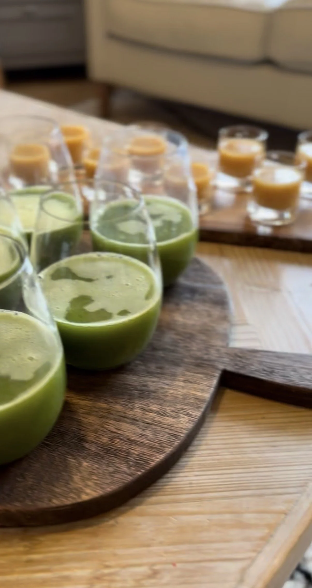 Close-up of a wooden tray with six glasses of green smoothie in the foreground and several small glasses of orange-colored beverage in the background, set on a wooden table.