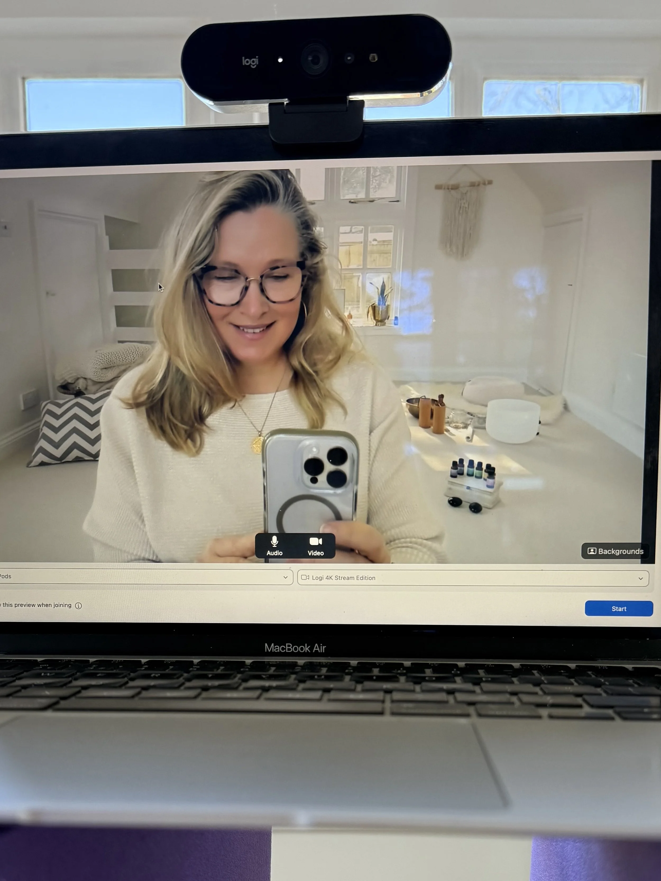 A woman with blonde hair and glasses taking a selfie with her phone in front of a mirror in a bright room, reflected on a computer screen.
