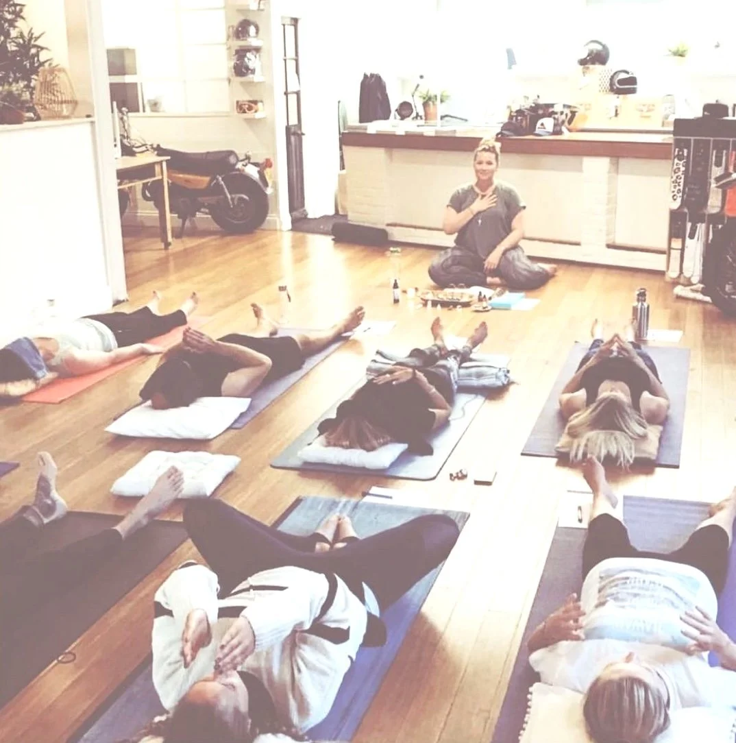 People participating in a yoga class inside a room with wooden floors and bright lighting, lying on yoga mats in a relaxed pose.