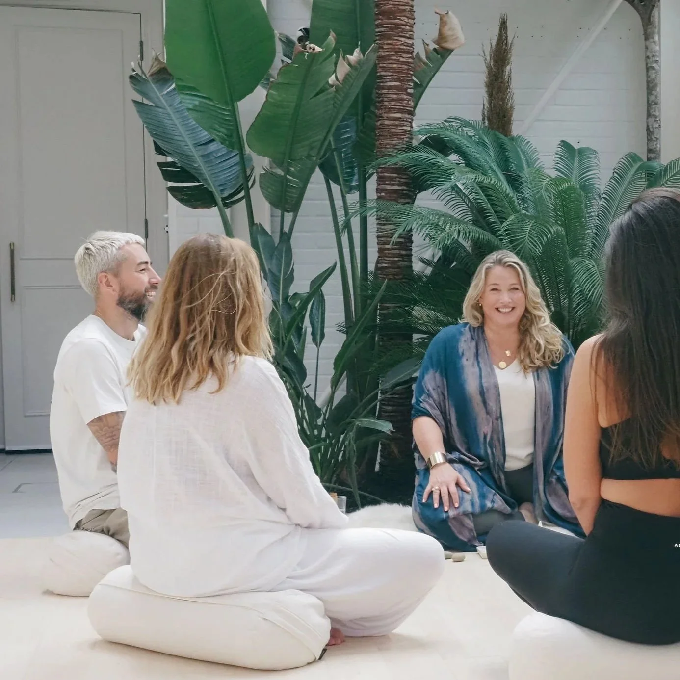 A group of five people sitting on the floor, smiling and having a conversation in a room with large green tropical plants.