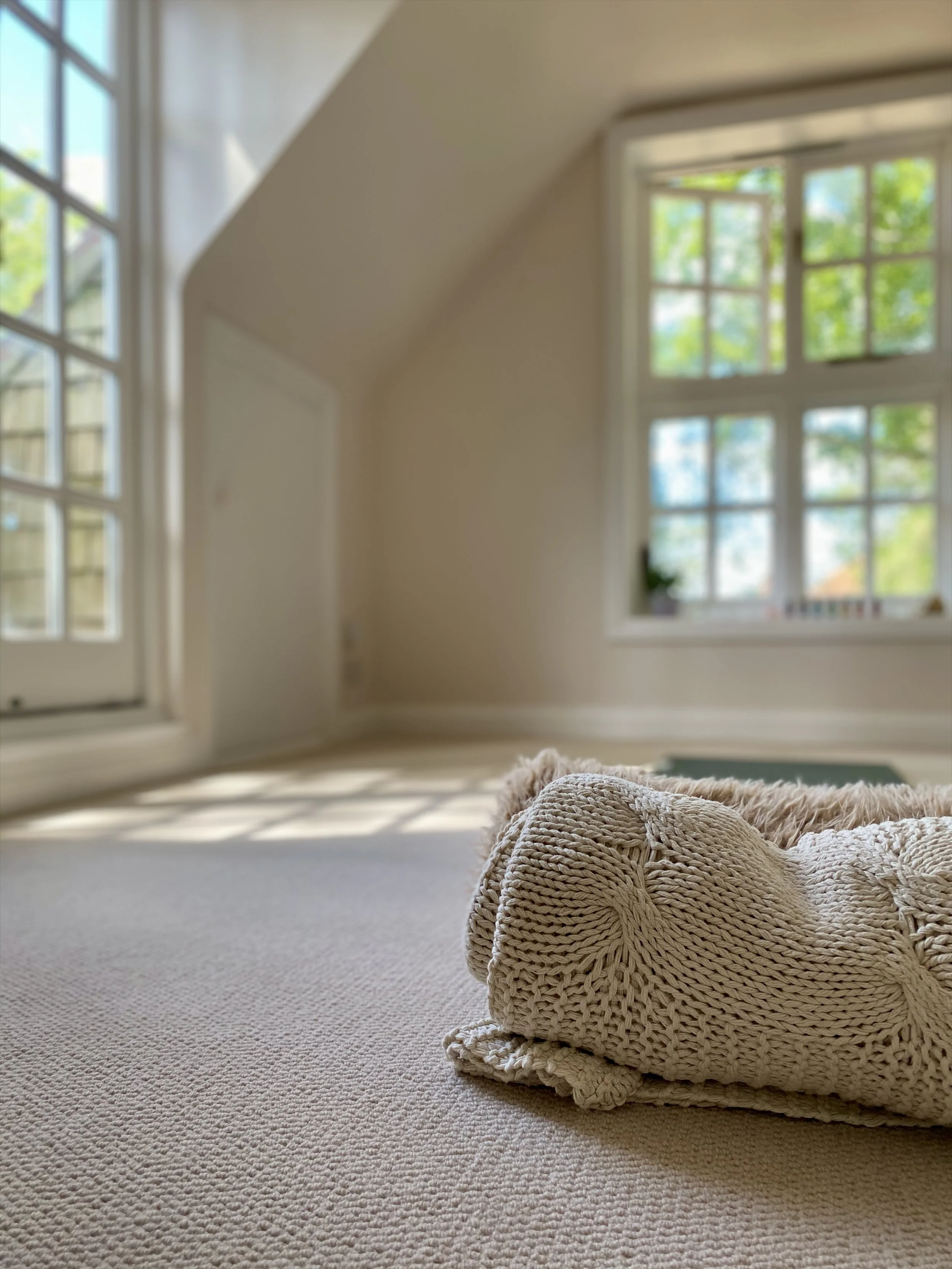 A cream-colored knitted blanket or pillow on a light beige carpet in a bright room with large windows showing green trees outside.