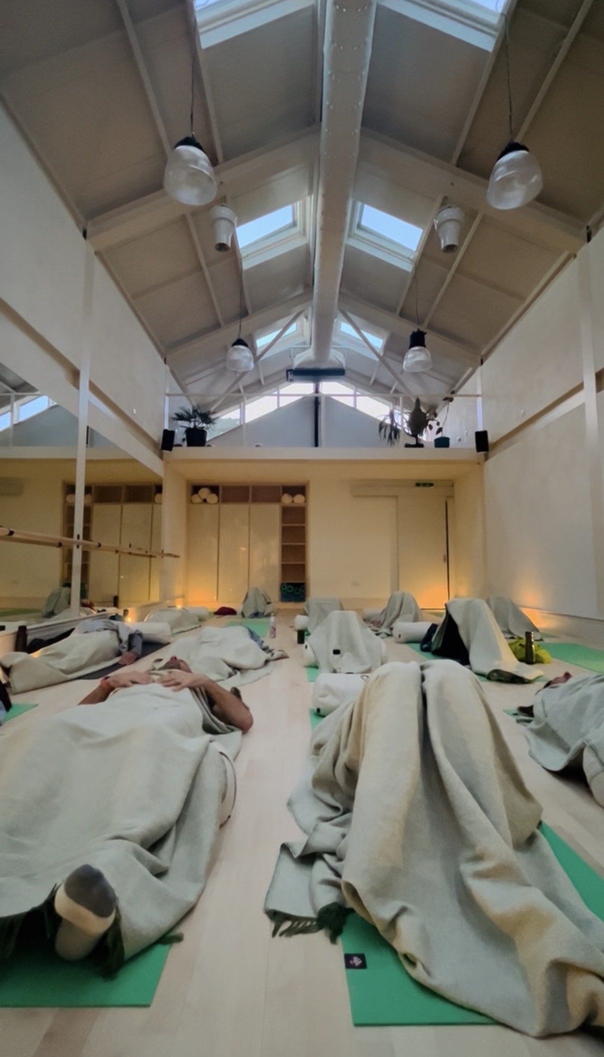 A yoga or meditation class with people lying on mats covered with blankets, in a spacious room with high ceilings, large windows, and skylights.