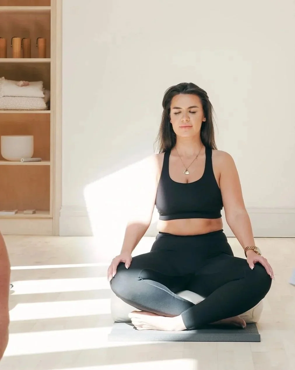 A woman practicing yoga indoors, sitting cross-legged on a yoga mat with her eyes closed in meditation.