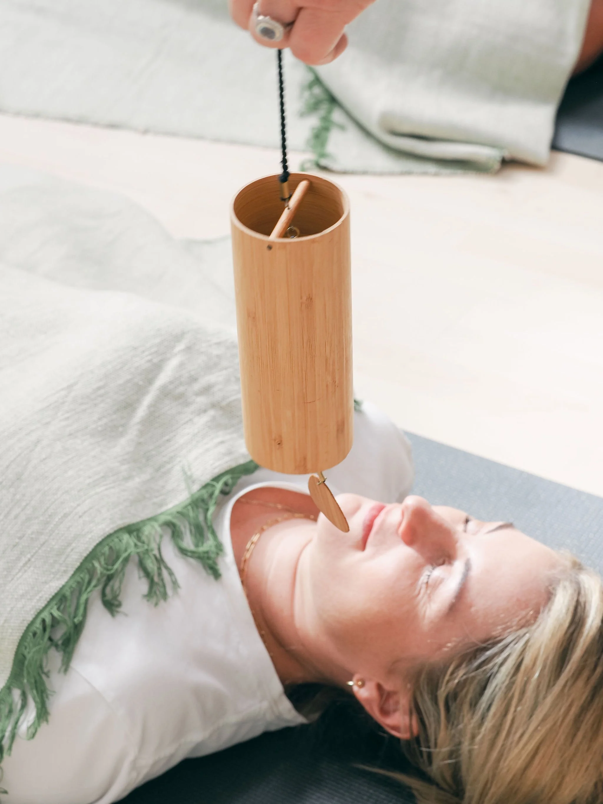 A person lying down with their eyes closed, holding a wooden suncare or sauna stick, dressed in white with green embroidery, and wearing eye makeup. The person has light-colored hair and is shown in a relaxing pose.